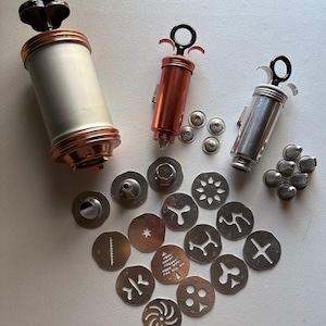 May include: A collection of cookie press tools including one large silver and copper-toned cylinder, two smaller handheld presses in copper and silver, and a variety of metal piping nozzles and flat patterned discs for shaping dough.