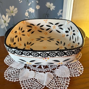 May include: A rectangular ceramic baking dish with black handles and a floral design in black and yellow. The dish sits on a white lace doily with heart-shaped cutouts. The rim of the dish is black.