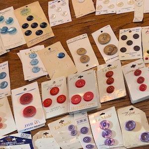 May include: A collection of various buttons on cards, in shades of blue, red, pink, brown, and purple. The buttons are in different sizes and styles, including round, flat, and domed. Some cards have text.