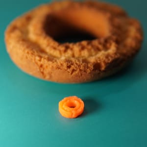 Puede incluir: Un pastel circular grande de color marrón dorado con una superficie texturizada y un agujero central, colocado detrás de un pequeño anillo de plástico naranja brillante que imita la forma del pastel sobre una superficie de color verde azulado.