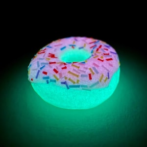Op de afbeelding: Een gloeiende donut met een roze glazuurlaag bedekt met kleurrijke hagelslag. De onderste helft van de donut gloeit felgroen, wat een speels effect creëert. De achtergrond is zwart.