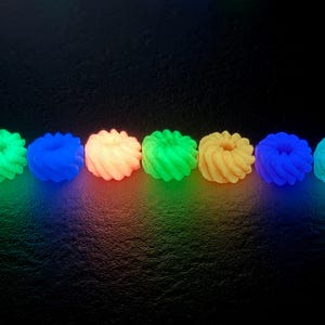 K&ouml;nnte beinhalten: Reihe von sechs leuchtenden, spiralf&ouml;rmigen Objekten in verschiedenen Farben: Gr&uuml;n, Blau, Orange und Gelb. Die Objekte sind gleichm&auml;&szlig;ig vor einem dunklen Hintergrund angeordnet, mit farbigem Licht, das von unten ausgeht.