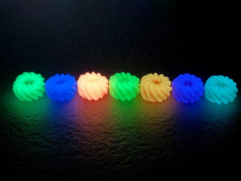 K&ouml;nnte beinhalten: Reihe von sechs leuchtenden, spiralf&ouml;rmigen Objekten in verschiedenen Farben: Gr&uuml;n, Blau, Orange und Gelb. Die Objekte sind gleichm&auml;&szlig;ig vor einem dunklen Hintergrund angeordnet, mit farbigem Licht, das von unten ausgeht.