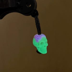 May include: A small skull-shaped bead featuring a glowing light green lower section and a purple top section, suspended from a black braided cord. The skull bead has detailed eye sockets, a nasal cavity, and a defined jawline, hanging vertically.