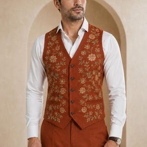 May include: A rust-coloured waistcoat featuring intricate gold floral embroidery, a V-neckline, and five dark buttons down the front. The waistcoat is worn over a crisp white long-sleeved button-down shirt, paired with matching rust-coloured trousers.