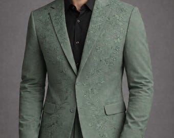 May include: Sage green suit with floral embroidery. The suit features a two-button jacket with notched lapels and a matching pair of trousers. A black button-down shirt is worn underneath the jacket. The suit is made of a soft, textured fabric.