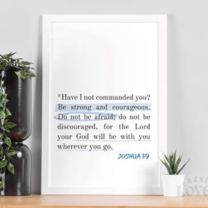 May include: A framed print featuring a passage from Joshua 1:9, with key phrases highlighted. The text is in a modern font, set against a white background. The print is displayed on a wooden surface, with a plant and decorative vase in the background.