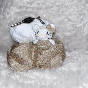 Peut inclure: Un porte-clés fait à la main en crochet représentant un chat blanc avec un nœud, assis sur une pelote de laine. Une clé argentée est à côté de la laine. La laine est blanche et dorée.