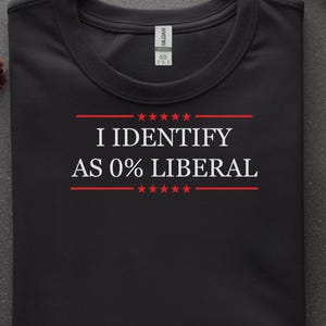 May include: Black t-shirt with the text "I IDENTIFY AS 0% LIBERAL" in white and red. The text is centered and framed by red lines and stars. The shirt is displayed on a dark gray surface with decorative fall elements.