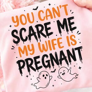 Pode incluir: Uma t-shirt rosa claro com o texto laranja e preto "YOU CAN'T SCARE ME MY WIFE IS PREGNANT" numa fonte de estilo escorrido. O design inclui pequenas ilustrações pretas de dois fantasmas, vários morcegos e pequenas estrelas espalhadas.