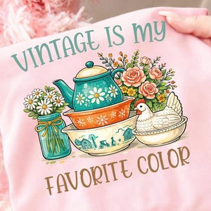 Puede incluir: Camiseta rosa claro con el texto "VINTAGE IS MY FAVORITE COLOR". El gráfico presenta una tetera turquesa, cuencos apilados, un tarro de margaritas, un ramo de rosas y una gallina en un cuenco. Diseño vintage y rústico.