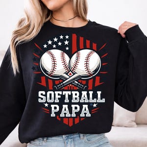 Op de afbeelding: Zwart sweatshirt met een afbeelding van twee gekruiste softbalballen, een rood-wit-blauw ontwerp en de tekst "SOFTBALL PAPA". De softbalballen zijn wit met rode stiksels.