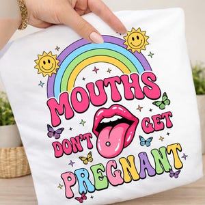 Peut inclure: Un t-shirt blanc avec un motif coloré comprenant un arc-en-ciel, deux soleils souriants et des papillons. Le texte "MOUTHS DON'T GET PREGNANT" est écrit en grandes lettres multicolores autour d'une illustration de bouche avec un piercing.