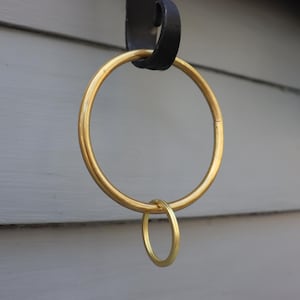 May include: A gold-coloured metal curtain ring with a smaller ring attached. The large ring is suspended from a black metal bracket. The background is a light grey wooden wall.