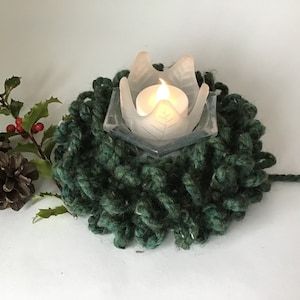May include: A handmade, dark green knitted wreath with a clear glass candle holder in the center. The candle holder has a leaf design and holds a lit candle. Pinecones and holly with red berries are on the left.