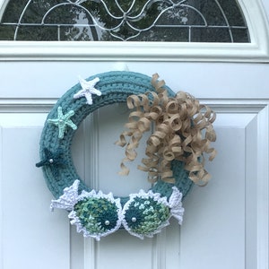 May include: A handmade teal crochet wreath decorated with white and pastel starfish, two crochet fish, and a tan paper ribbon. The wreath is displayed on a white door with a glass window.