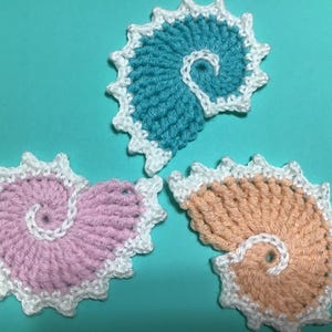 Könnte beinhalten: Drei gehäkelte Spiralformen in Rosa, Pfirsich und Türkis mit weißem Rand. Die Formen sind in einem Dreieck auf einem türkisfarbenen Hintergrund angeordnet.