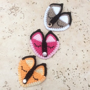 May include: Three crocheted fox faces with closed eyes and black button noses. The foxes are pink, orange, and brown and white.