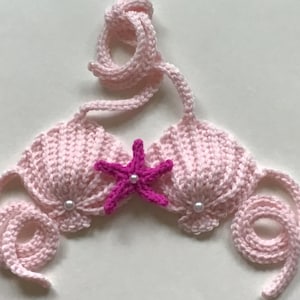 May include: A light pink crocheted bikini top featuring two shell-shaped cups with a small white pearl bead at the center of each. A bright fuchsia crocheted starfish is attached between the cups, and the piece includes long, curled pink ties.