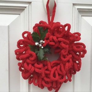 May include: A vibrant red loop yarn wreath, adorned with green holly leaves and silver berry decorations. This festive wreath hangs on a white door. The wreath is approximately 30 cm in diameter, ideal for seasonal decoration.