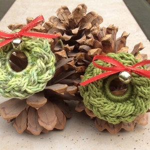 May include: Two green crocheted wreaths with red bows and gold bells, sitting on a brown pine cone.