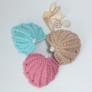May include: Three crocheted seashells in pink, blue, and brown with pearl accents. The shells are arranged in a triangle shape on a white background.