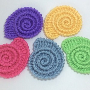 Puede incluir: Cinco conchas de mar de ganchillo en diferentes colores: morado, amarillo, rosa, azul y verde. Las conchas son todas del mismo tamaño y forma, y están dispuestas en círculo.
