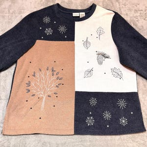 Pode incluir: Uma camisola de manga comprida com um design em blocos de cores. A camisola apresenta painéis azul marinho, pêssego e branco. É adornada com motivos bordados com temas de inverno e outono, incluindo flocos de neve, folhas e uma árvore.