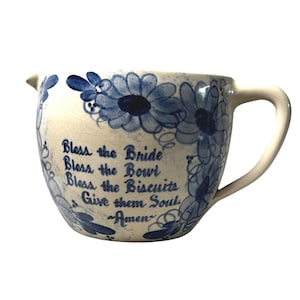 May include: A cream-colored ceramic mixing bowl with a handle and a pouring spout, featuring blue hand-painted floral patterns and the text: Bless the Bride, Bless the Bowl, Bless the Biscuits, Give them Soul, ~Amen~. The surface has a crackled finish.