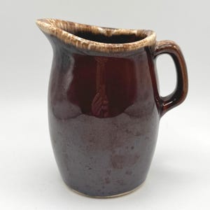 May include: A dark brown ceramic pitcher with a glossy finish and a lighter cream-colored drip glaze along the rim. The pitcher features a curved handle on the right side and a wide pouring spout, showcasing a smooth, rounded body design.