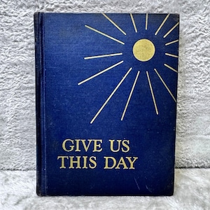 May include: A vintage book with a navy blue cover featuring a gold sunburst design. The title "GIVE US THIS DAY" is printed in gold lettering. The book has a worn appearance, suggesting it is an antique.