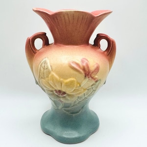 May include: A ceramic vase featuring a gradient color scheme that transitions from a soft pink at the flared rim to a warm yellow in the center and a muted teal at the base. The body is decorated with a raised floral relief and two side handles.