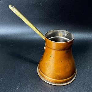 May include: A copper-colored metal coffee pot with a wide, rounded base and a narrow neck. It features a long, slender, metallic handle extending diagonally from the side and a small pouring spout integrated into the top rim of the vessel.