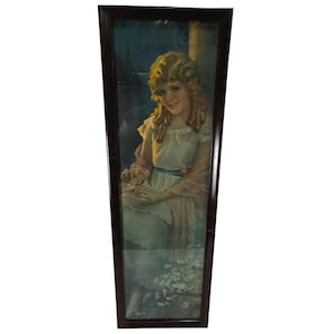 May include: A vertical rectangular framed print featuring a portrait of a person with long blonde curly hair wearing a white dress with a blue sash and a light pink shawl. The person is seated outdoors among white flowers with a dark background.