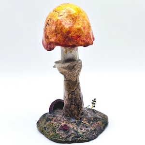 Può includere: Una scultura a forma di fungo realizzata a mano con un cappello strutturato arancione e giallo e un gambo beige. La base è screziata di verde e marrone, con una piccola apertura scura e un rametto di vegetazione. La scultura è su sfondo bianco.