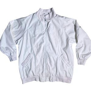 May include: A light-colored, zip-up jacket with a ribbed collar, cuffs, and hem. The jacket has two front pockets and a full-length zipper. The material appears to be a lightweight fabric, suitable for layering. The jacket is a neutral color.
