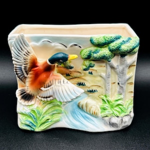 May include: A rectangular ceramic planter featuring a three-dimensional mallard duck in flight with a green head, orange chest, and white wings. The background depicts a painted landscape with green trees, a blue stream, and light yellow sky.