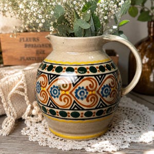 May include: A beige ceramic pitcher featuring a hand-painted pattern of dark green circles, yellow wavy lines, and orange scrollwork with blue floral accents. The pitcher has a rounded body, a wide handle, and a small spout at the top rim.