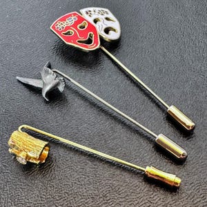 May include: Three decorative lapel pins. One pin features red and white theatre masks. Another pin has a silver bird design. The third pin has a gold-coloured, textured cylindrical design with a small crystal.