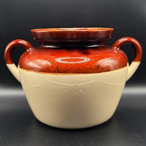May include: A two-handled ceramic crock featuring a glossy dark brown glazed upper section and a smooth cream-colored lower body. The vessel has a wide, rounded opening with a thick rim and two curved handles extending from the sides.