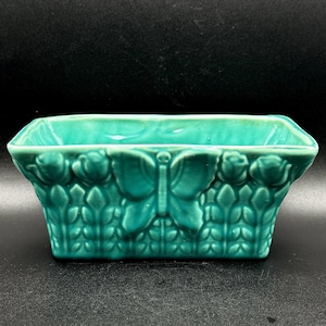 May include: A rectangular teal ceramic planter featuring a raised relief design of a central butterfly flanked by rose buds and a lattice pattern. The vessel has a glossy finish and a wide, open top with a flared rim, showcasing a consistent monochromatic color throughout the entire piece.