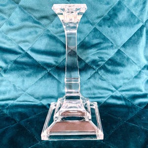 Può includere: Un portacandele in vetro trasparente ed elegante, con base quadrata e stelo alto e sottile. La parte superiore è progettata per contenere una singola candela. Il portacandele è alto circa 25 cm, perfetto per aggiungere un tocco di raffinatezza.