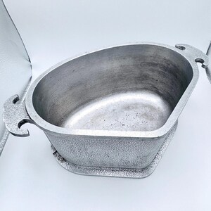 Puede incluir: Una fuente de metal plateado de forma ovalada con asas decorativas. La fuente tiene un exterior texturizado y se asienta sobre una base a juego. El interior de la fuente es liso y muestra signos de uso.
