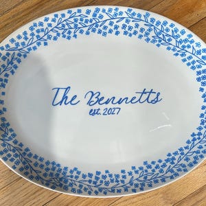 May include: An oval white ceramic serving platter featuring a blue floral vine border pattern. The center of the platter displays the blue text The Bennetts and est. 2027 in a script and sans-serif font combination, creating a personalized design.