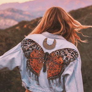 May include: A light blue denim jacket with a beaded butterfly design on the back. The butterfly is orange, black, and iridescent.