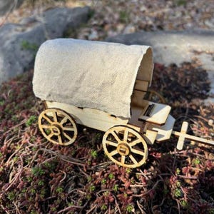 May include: A miniature wooden covered wagon with a canvas top. The wagon features intricate laser-cut details on the wheels and frame. The wagon is set against a backdrop of greenery and rocks, suggesting an outdoor setting.