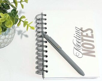 May include: A gray pen rests on a white spiral-bound notebook with the words "Meeting NOTES" printed on the cover. A small green plant in a decorative glass vase is in the upper left corner. The pen is angled across the notebook.