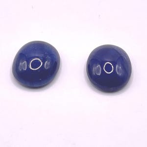 May include: Two oval-shaped, dark blue glass beads. Each bead has a glossy finish and a small, circular indentation on one side. The beads are displayed against a white background.