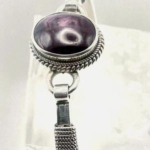 May include: A silver cuff bracelet featuring a central oval-shaped purple stone with dark swirling patterns. The stone is set in a twisted rope-style silver bezel, connected to a band with a textured, braided silver wire design on the sides.