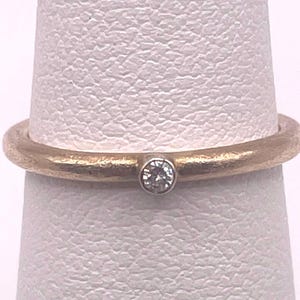 May include: A gold ring with a textured, matte finish featuring a single, small, round, clear gemstone set in a silver-toned bezel at the center. The band has a consistent thickness and a rounded profile, showcasing a subtle contrast between the gold metal and the silver setting.
